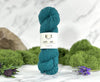 Teal skein of yarn with a label on a white surface with greenery in the background