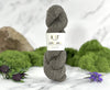A skein of gray yarn standing upright on a white marble surface, wrapped with a white paper label that reads 'KIT Low Amano.' Green moss, purple flowers, and a rock are arranged around it as styled props.