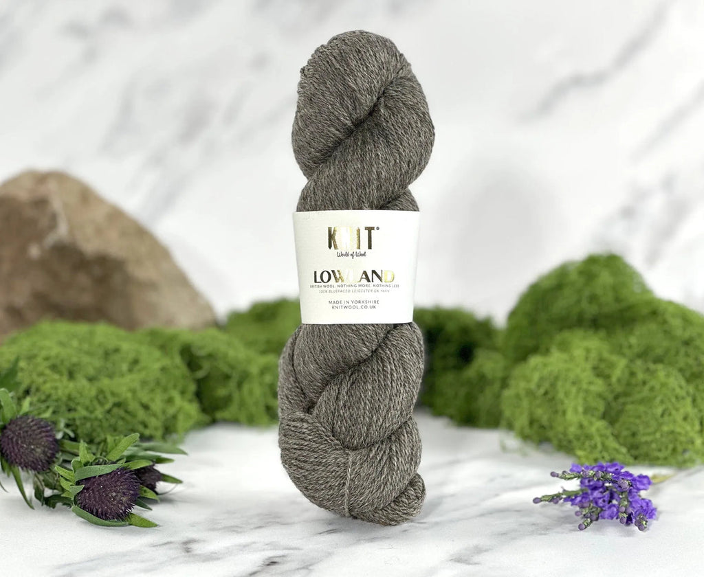 A skein of gray yarn standing upright on a white marble surface, wrapped with a white paper label that reads 'KIT Low Amano.' Green moss, purple flowers, and a rock are arranged around it as styled props.