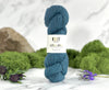 Blue skein of yarn with a label on a marble surface surrounded by greenery and flowers.