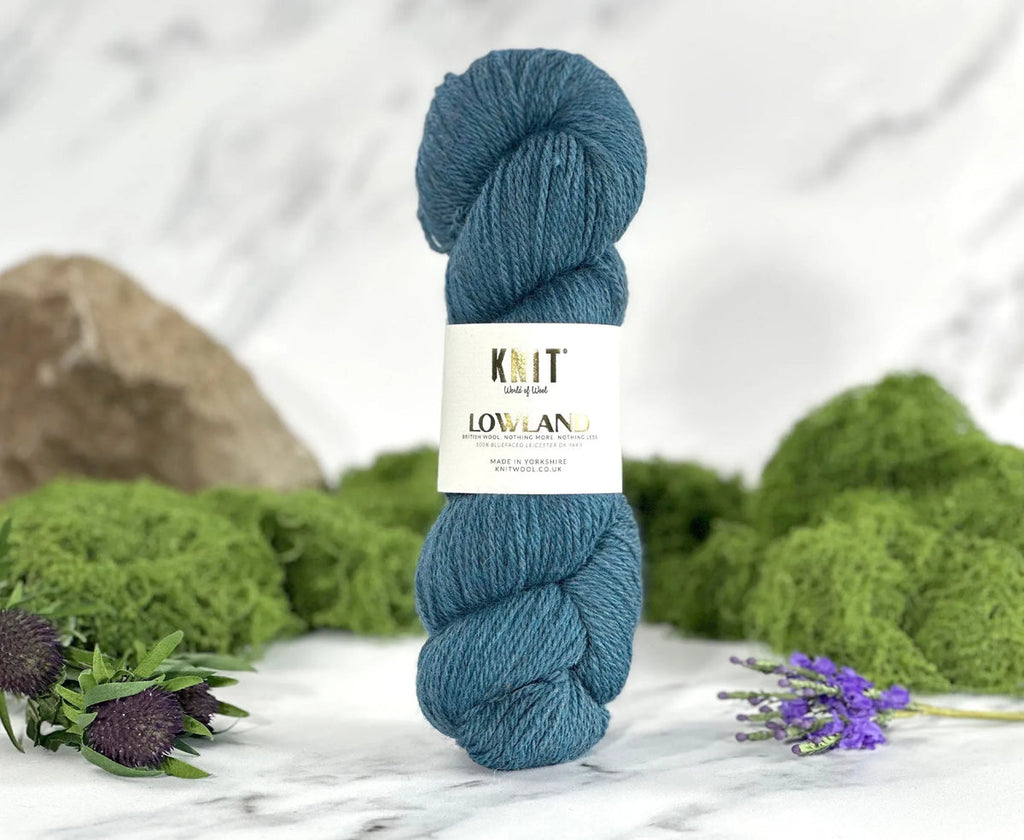 Blue skein of yarn with a label on a marble surface surrounded by greenery and flowers.