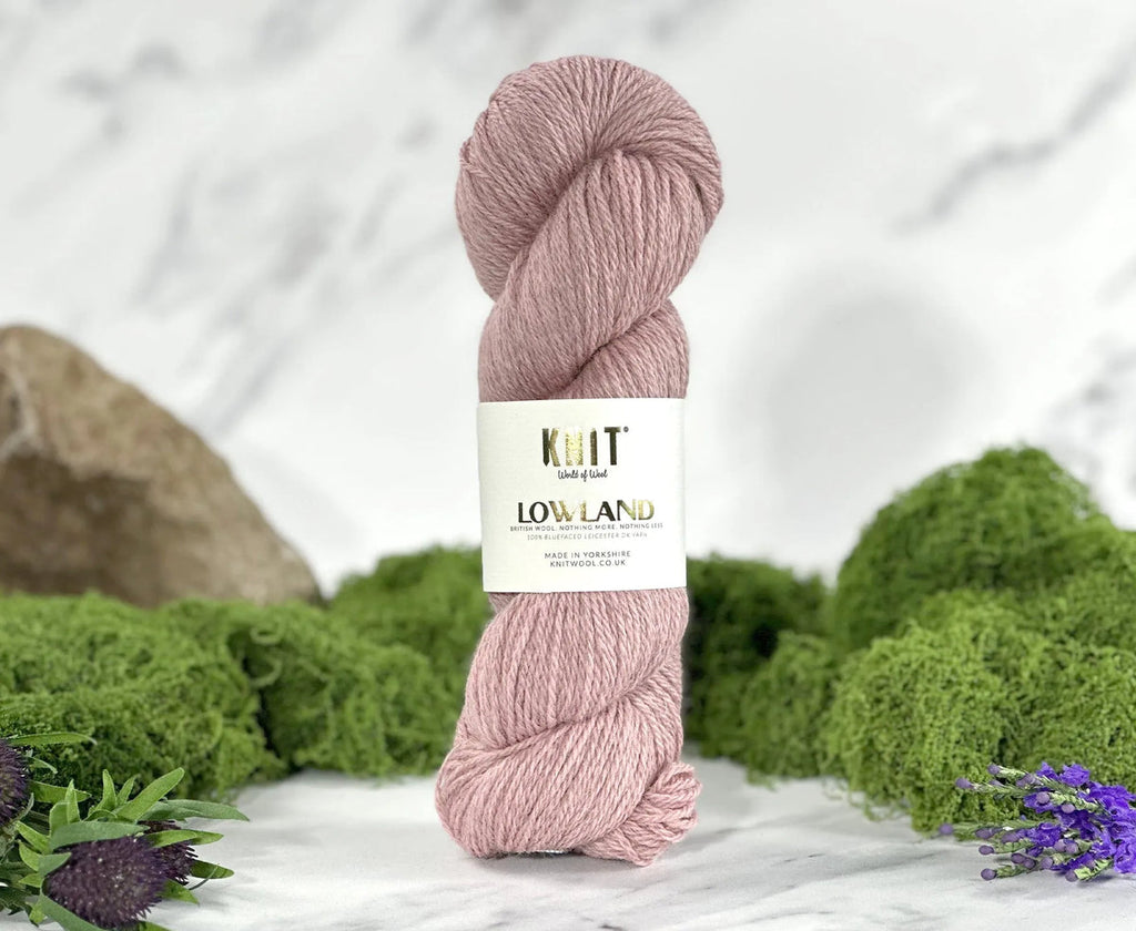 A skein of dusty pink yarn from Knit Low Land, wrapped in a white paper label, set against a blurred background of green moss, purple flowers, and a light marble surface.