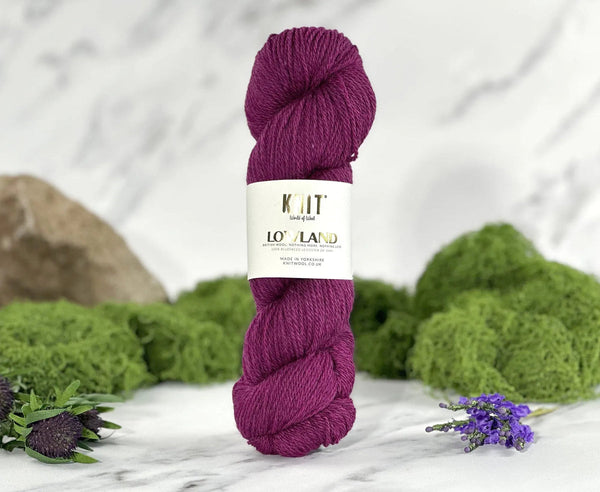 A skein of deep purple yarn with a white paper label reading 'Knit Loveland' and '100% Wool', styled on a white marble surface with green moss, a rock, and small purple flowers.