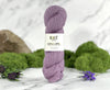 A skein of lavender purple yarn standing upright on a white marble surface, with a white label reading 'KNIT LOWLAND'. Green moss, purple flowers, and a blurred stone are arranged around it against a soft white marble background.