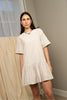 A woman wearing a short-sleeve, off-white tiered tunic dress, standing in a neutral-toned indoor setting with draped fabric in the background.