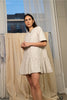 Woman wearing a beige dress standing in a room with curtains and a window.