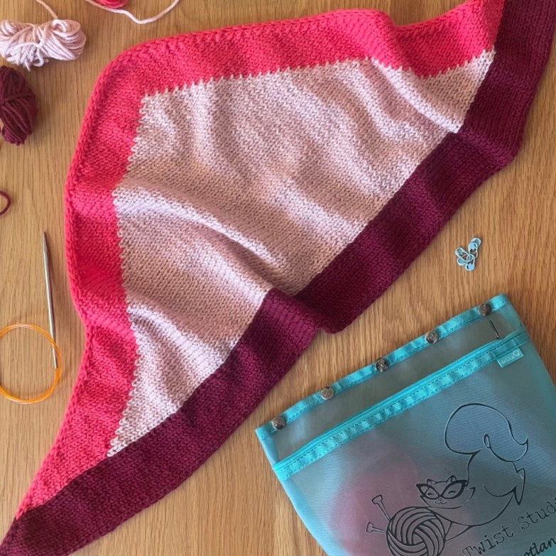 Knitted fabric with red and pink stripes on a wooden surface with yarn and a blue pouch.