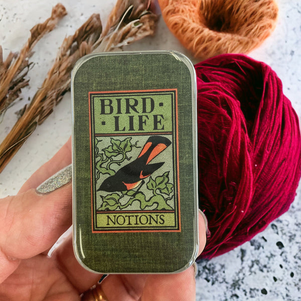 Hand holding a green tin labeled 'Bird Life Notions' with yarn and dried plants in the background.