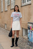 A woman stands on a cobblestone street wearing a light gray graphic t-shirt with 'NEW YORK' in red and blue, a white mini skirt, and black knee-high boots, holding a black tote bag.