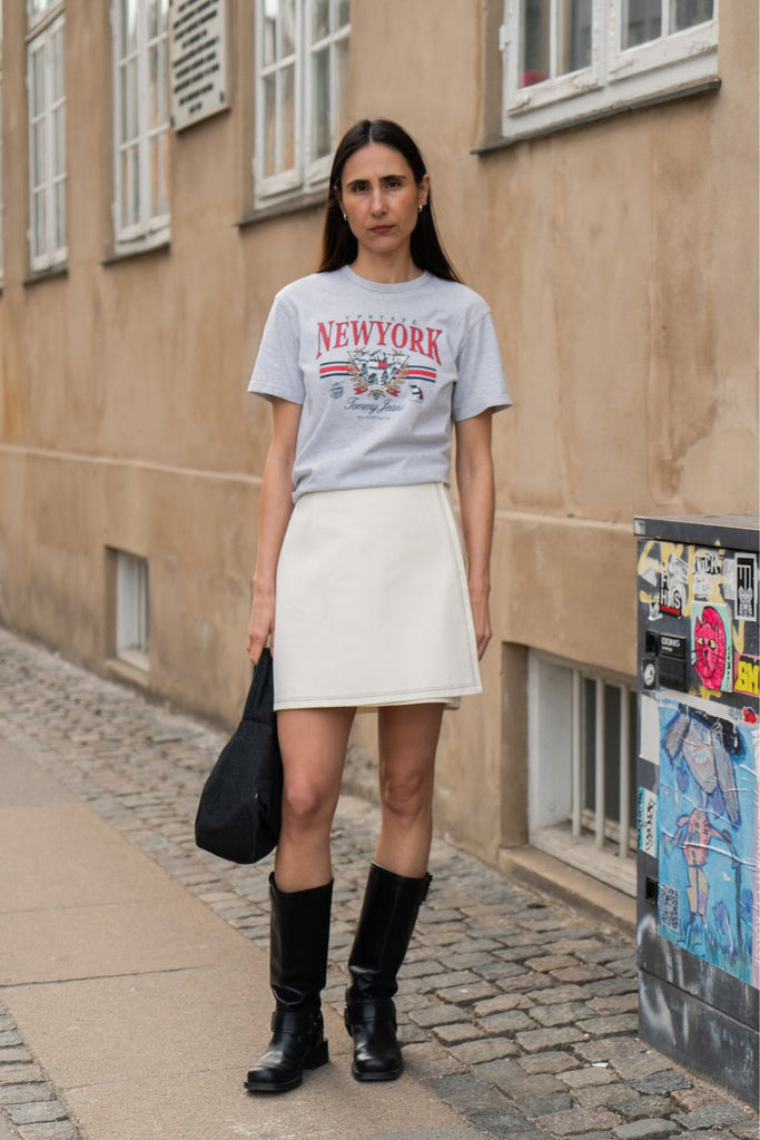 A woman stands on a cobblestone street wearing a light gray graphic t-shirt with 'NEW YORK' in red and blue, a white mini skirt, and black knee-high boots, holding a black tote bag.