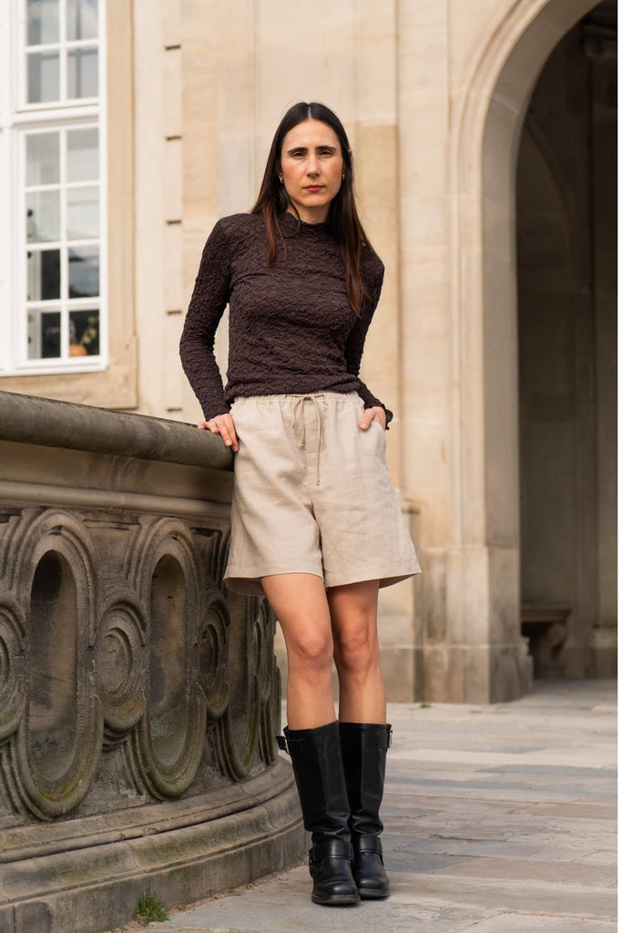 Woman standing outdoors in front of a stone building wearing a brown top, beige shorts, and black boots.