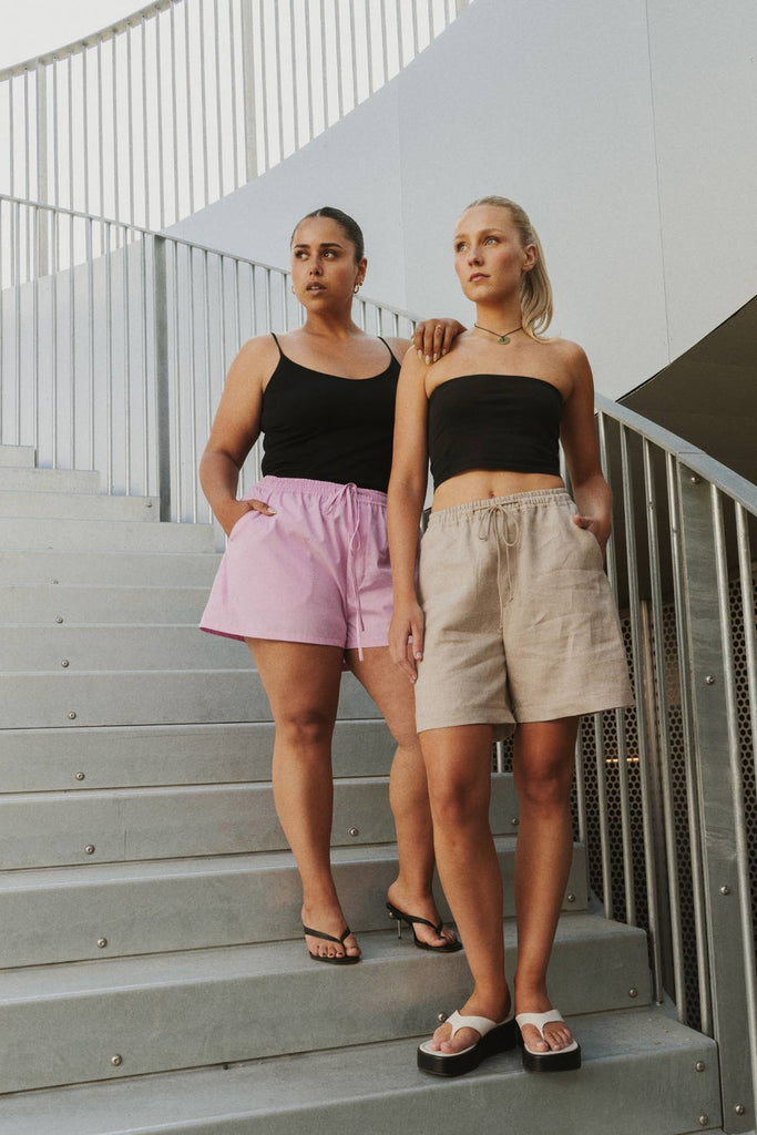 Two women standing on outdoor concrete stairs wearing matching black strapless crop tops and shorts in different colors: one in light pink shorts and the other in beige shorts.