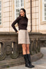 A woman stands outdoors in front of a stone building, wearing a dark brown textured long-sleeve top, light beige shorts, and black knee-high boots.