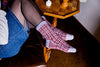 Person wearing checkered gloves and blue jeans sitting on a chair with a wooden table and chessboard in the background.