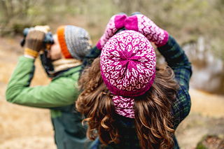 Two people in winter clothing looking through binoculars by a body of water.