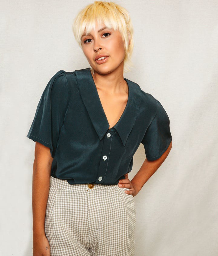 Person wearing a dark green blouse with a collar and checkered pants against a plain background