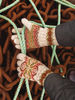 A close-up of a person's hands wearing cream knit gloves with a colorful red, green, and orange Fair Isle pattern, holding onto thick green and white ropes over a bed of rusty brown chains.