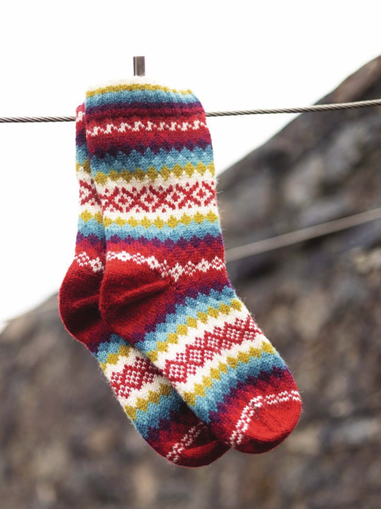 A pair of hand-knit socks with a colorful Fair Isle pattern, hanging from a wire rope outdoors against a blurred rocky background.