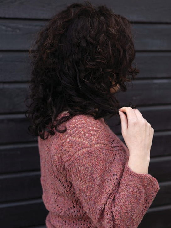 A person shown from the back wearing a dusty rose knit sweater, with dark curly hair, standing against a dark horizontal wood plank background.