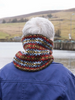 A person viewed from behind wearing a colorful knitted cowl neck warmer with a red, blue, yellow, and black pattern, standing outdoors near water and hills.
