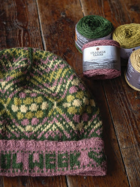 A close-up of a hand-knit beanie hat with a green, pink, and cream Fair Isle pattern, laid on a rustic wooden table next to several skeins of yarn, including a visible label for 'URABALE YARNS'.