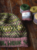 A close-up of a hand-knit beanie hat with a green, pink, and cream Fair Isle pattern, laid on a rustic wooden table next to several skeins of yarn, including a visible label for 'URABALE YARNS'.