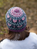 A person viewed from behind wearing a knitted beanie hat with a pink, purple, and gray fair isle pattern, set against a blurred outdoor background.