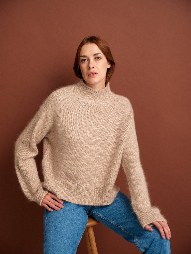 Woman wearing a beige sweater and blue jeans sitting on a stool against a brown background