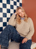 Person wearing a beige sweater and blue jeans sitting against a checkered wall.