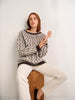 Woman wearing a patterned sweater and white pants sitting on a wooden block against a white wall.