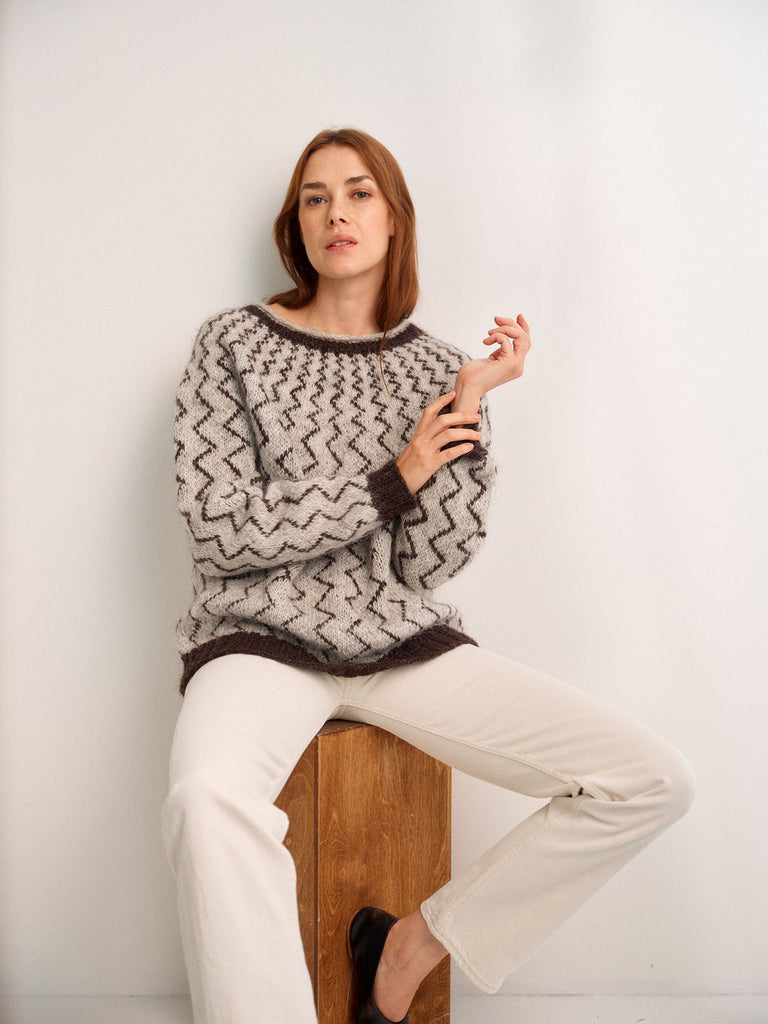 Woman wearing a patterned sweater and white pants sitting on a wooden block against a white wall.