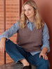 Woman wearing a brown knitted vest over a blue shirt and jeans, sitting against a wooden wall.