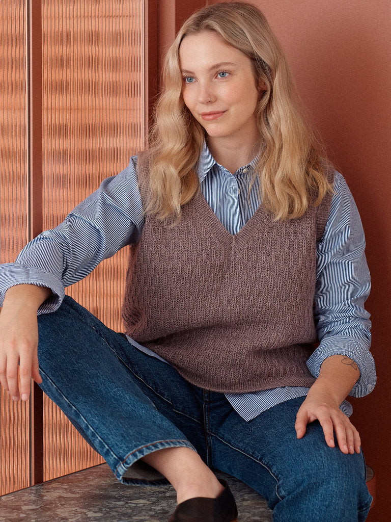 Woman wearing a brown knitted vest over a blue shirt and jeans, sitting against a wooden wall.