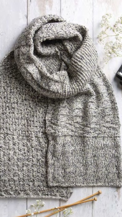 Knitted gray scarf on a wooden surface with knitting needles and flowers.
