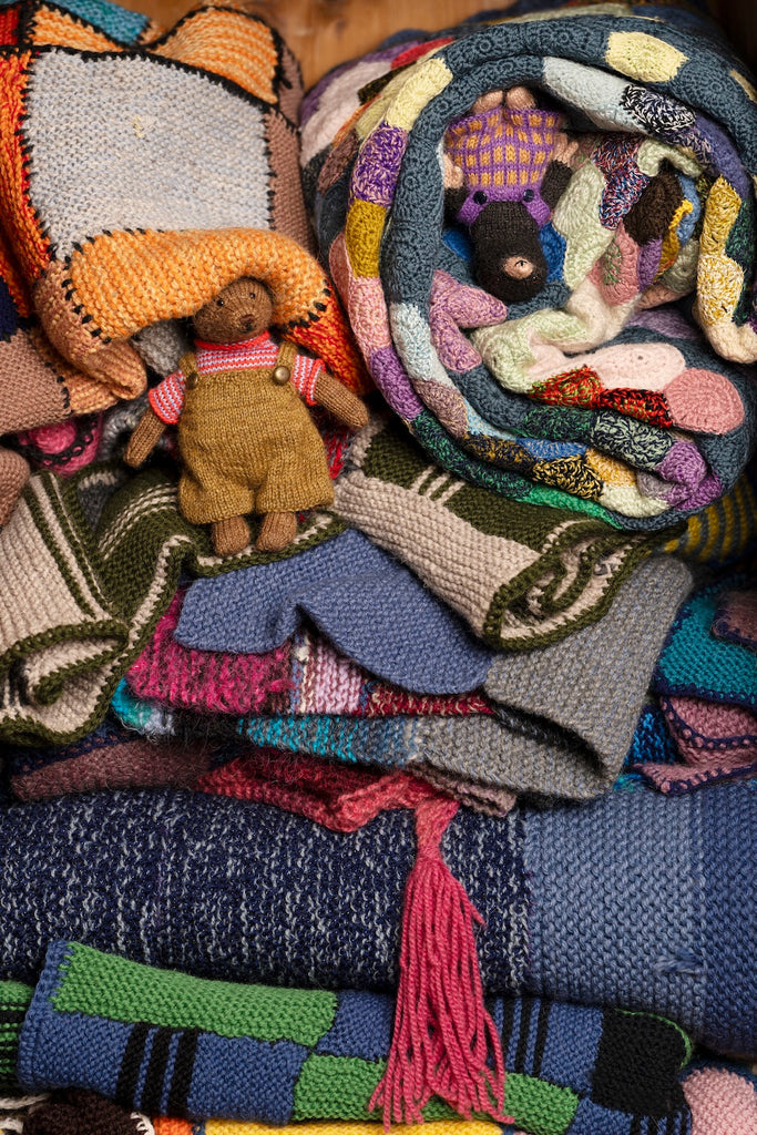 A close-up of a colorful assortment of knitted blankets, scarves, and woven textiles piled together, with a small plush teddy bear doll nestled inside the stack.