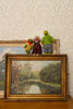 A framed landscape painting of a river scene is displayed on a wooden surface against patterned wallpaper, with three small plush stuffed animals resting on top of the frame.
