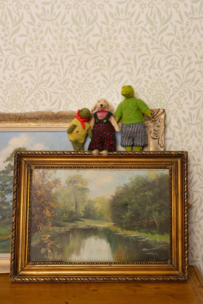 A framed landscape painting of a river scene is displayed on a wooden surface against patterned wallpaper, with three small plush stuffed animals resting on top of the frame.