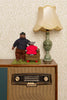 A styled tabletop scene with a vintage radio cabinet, a cream lamp, and two plush bear figures standing beside an armchair. The background is patterned wallpaper, and no visible brand markings appear on the products.