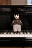 A small knitted badger toy wearing a brown vest with yellow buttons and patterned trousers, standing on the piano keys in front of a black grand piano.