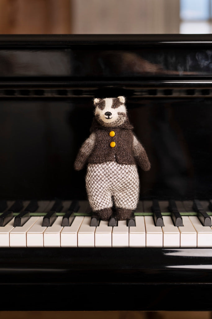 A small knitted badger toy wearing a brown vest with yellow buttons and patterned trousers, standing on the piano keys in front of a black grand piano.
