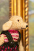 A close-up of a knitted plush dog toy wearing a pink and cream striped sweater and dark green overalls, positioned in front of a blurred golden frame.