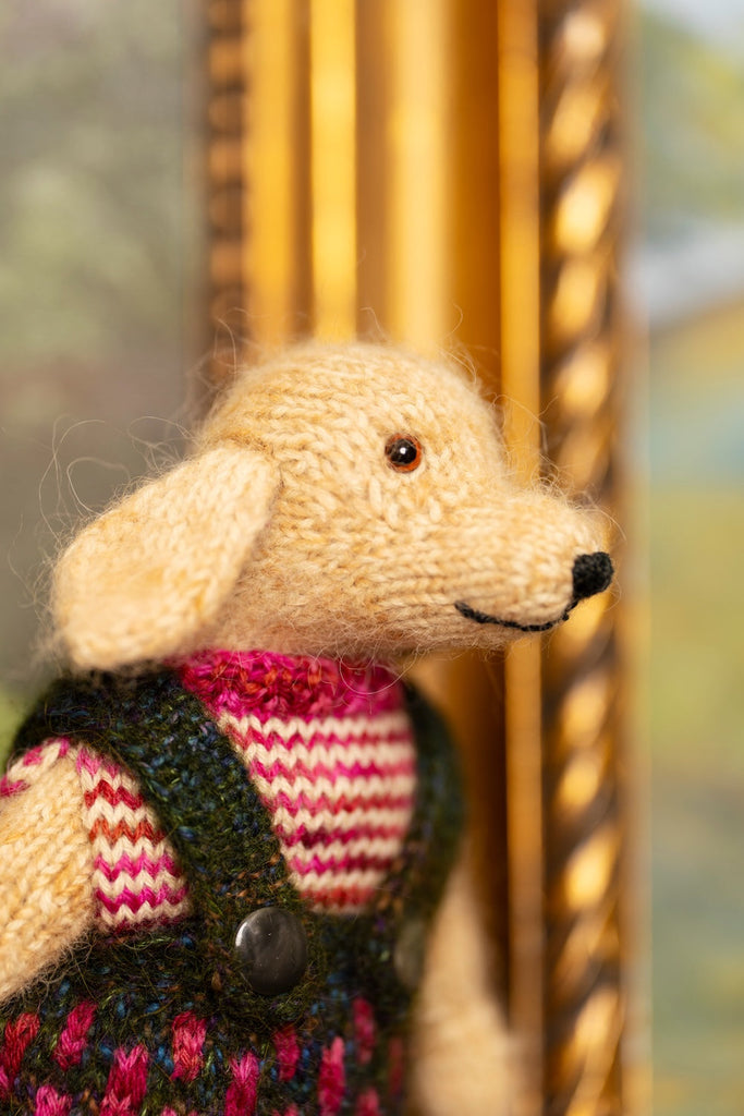 A close-up of a knitted plush dog toy wearing a pink and cream striped sweater and dark green overalls, positioned in front of a blurred golden frame.