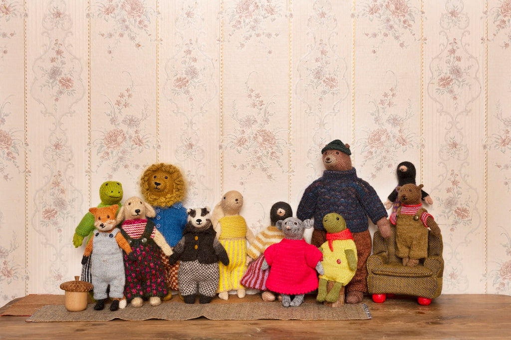 A group of handmade plush teddy bear figures and animal characters posed together on a wooden surface against a vintage floral wallpaper background.