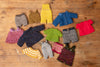 A collection of hand-knit doll clothes in various colors and patterns, including sweaters, shorts, and cardigans, arranged on a rustic wooden surface.
