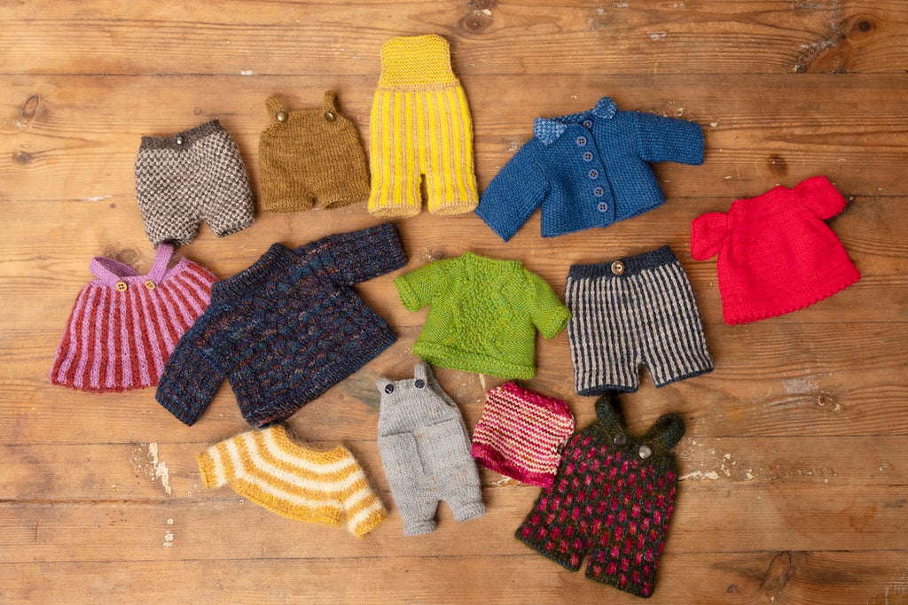 A collection of hand-knit doll clothes in various colors and patterns, including sweaters, shorts, and cardigans, arranged on a rustic wooden surface.