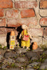 A small knitted rabbit doll wearing a yellow striped dress and holding a red-and-white checkered bag, posed outdoors against a brick wall on stones with toy blocks nearby.