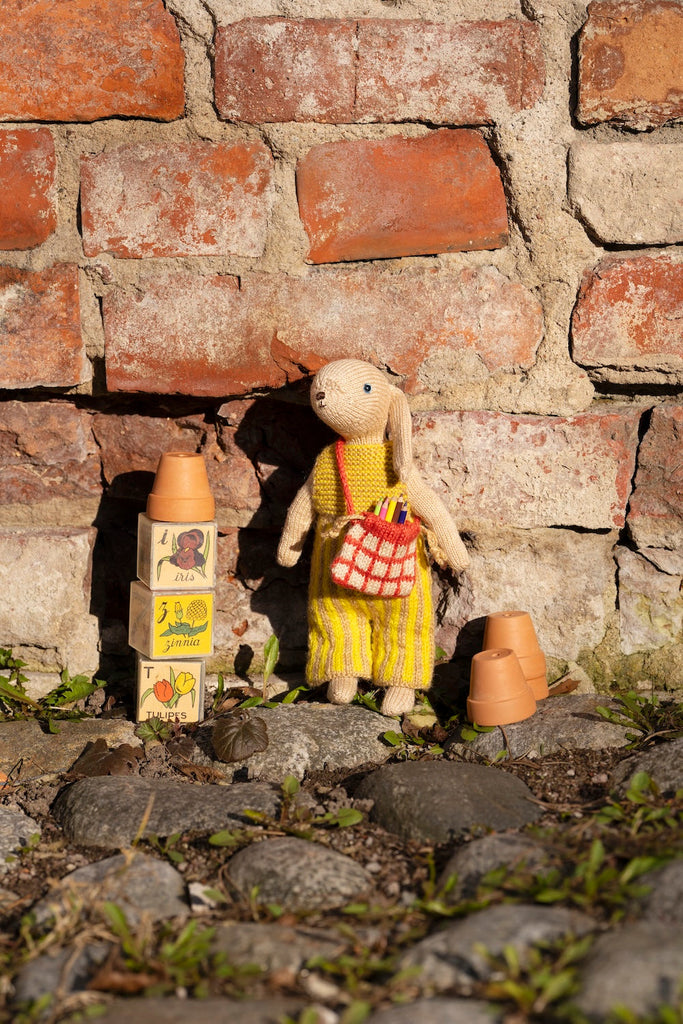 A small knitted rabbit doll wearing a yellow striped dress and holding a red-and-white checkered bag, posed outdoors against a brick wall on stones with toy blocks nearby.