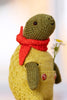 A knitted plush turtle toy wearing a red knit scarf and a mustard yellow sweater, holding a small white and yellow flower against a soft, out-of-focus light background.