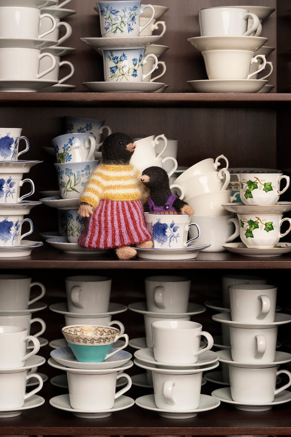 A collection of stacked white ceramic teacups and saucers on dark wooden shelves, with two small plush toy birds placed among the cups.