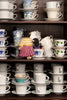 A collection of stacked white ceramic teacups and saucers on dark wooden shelves, with two small plush toy birds placed among the cups.